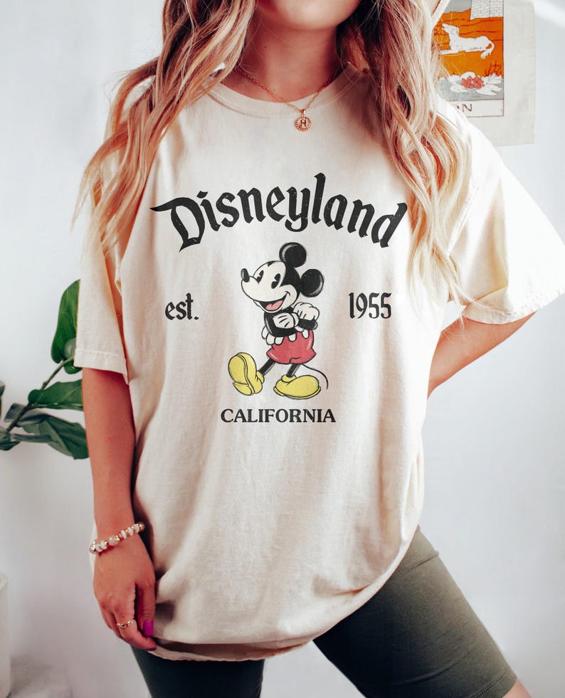 以下が含まれることがあります： クリーム色のTシャツに、ミッキーマウスの白黒のグラフィックがプリントされています。Tシャツには「Disneyland est. 1955 California」という文字がプリントされています。