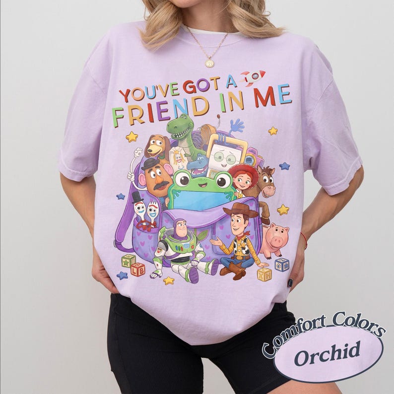 May include: A light purple t-shirt featuring the text "YOU'VE GOT A FRIEND IN ME" and a colorful graphic of various animated characters including a cowboy, a space ranger, a dinosaur, a potato figure, and a pig emerging from a purple backpack.