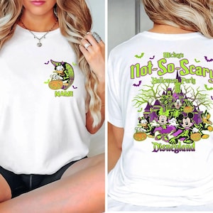 May include: White t-shirt featuring a Halloween-themed design. The front displays a cartoon character with a witch hat and a pumpkin. The back features the text "Mickey's Not-So-Scary Halloween Party" with Disney characters and a haunted castle.