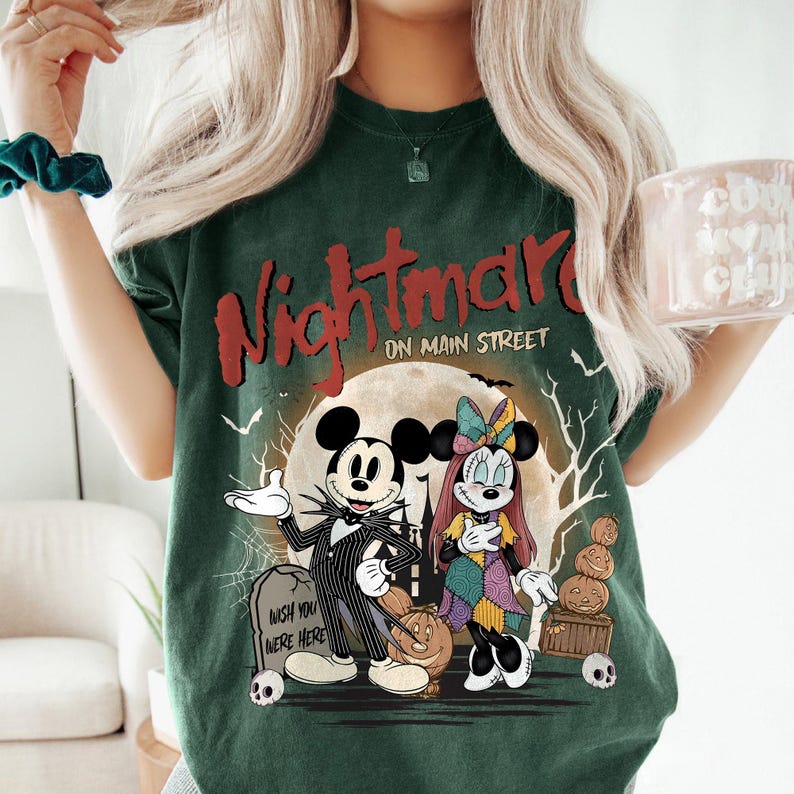 May include: Dark green t-shirt featuring a graphic from "The Nightmare Before Christmas" with Mickey Mouse and Minnie Mouse characters. The design includes the text "Nightmare on Main Street" and a spooky Halloween theme.
