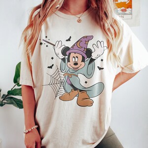 May include: Cream-colored t-shirt featuring a cartoon Mickey Mouse dressed as a wizard for Halloween. Mickey is wearing a purple wizard hat with a crescent moon, a blue robe, and brown shoes. Bats and a spiderweb are also featured in the design.