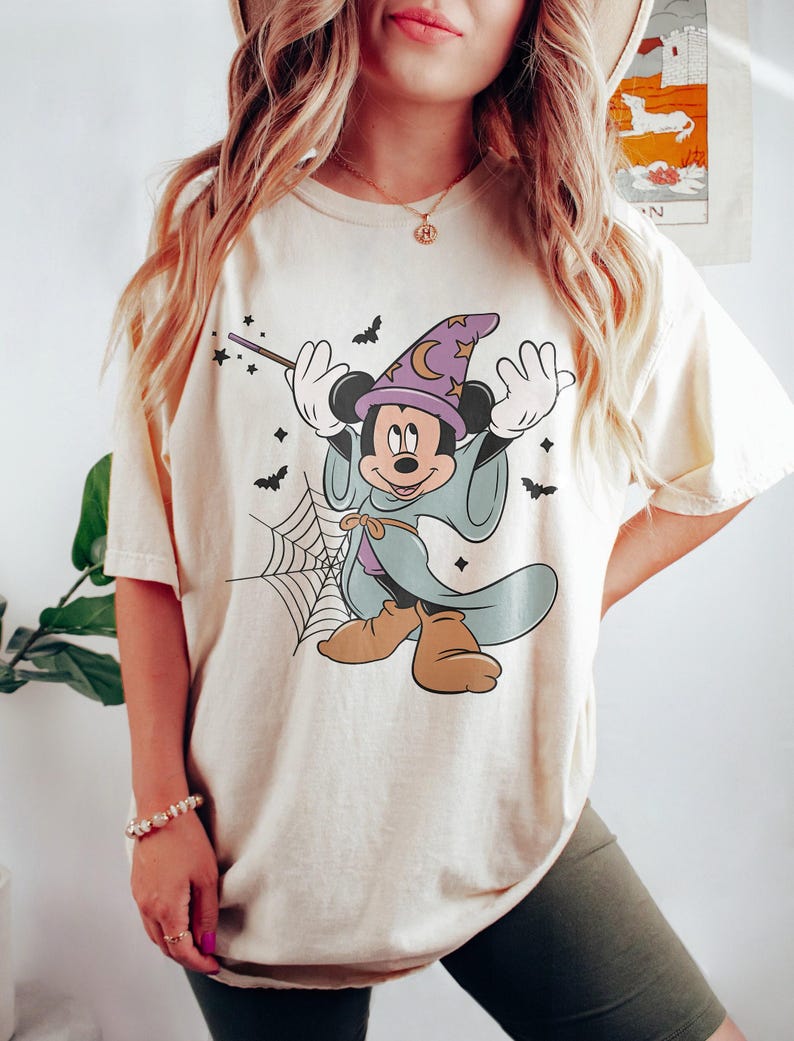 May include: Cream-colored t-shirt featuring a cartoon Mickey Mouse dressed as a wizard for Halloween. Mickey is wearing a purple wizard hat with a crescent moon, a blue robe, and brown shoes. Bats and a spiderweb are also featured in the design.
