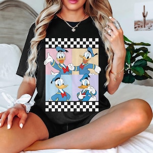 May include: Black t-shirt featuring a four-panel graphic of Donald Duck in various poses. The cartoon character is wearing a blue sailor suit and a red bow tie. The graphic is framed by a black and white checkered border.