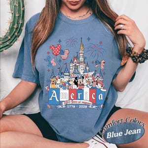 May include: A blue denim-colored t-shirt featuring a graphic print of a castle, fireworks, and cartoon characters wearing patriotic red, white, and blue outfits. The text reads America, 250 Years of Freedom, and 1776-2026 in a decorative font.