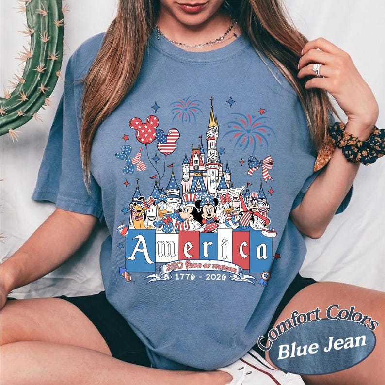 May include: A blue denim-colored t-shirt featuring a graphic print of a castle, fireworks, and cartoon characters wearing patriotic red, white, and blue outfits. The text reads America, 250 Years of Freedom, and 1776-2026 in a decorative font.
