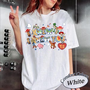 May include: A white t-shirt featuring a colorful graphic print of Toy Story characters including Woody, Buzz Lightyear, Jessie, Rex, and Hamm. The text reads I KNEW IT I KNEW YOU with a red heart containing the letters TS. The design includes small stars.