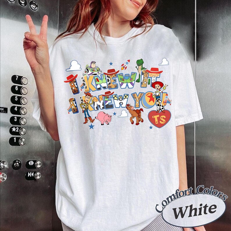 May include: A white t-shirt featuring a colorful graphic print of Toy Story characters including Woody, Buzz Lightyear, Jessie, Rex, and Hamm. The text reads I KNEW IT I KNEW YOU with a red heart containing the letters TS. The design includes small stars.