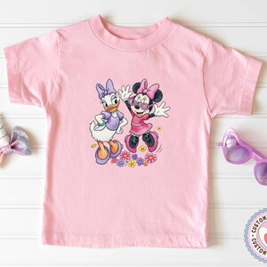 May include: A light pink t-shirt featuring a colorful graphic of Daisy Duck and Minnie Mouse. Daisy Duck is wearing a purple dress and Minnie Mouse is wearing a pink dress and sunglasses. The graphic includes colorful flowers.