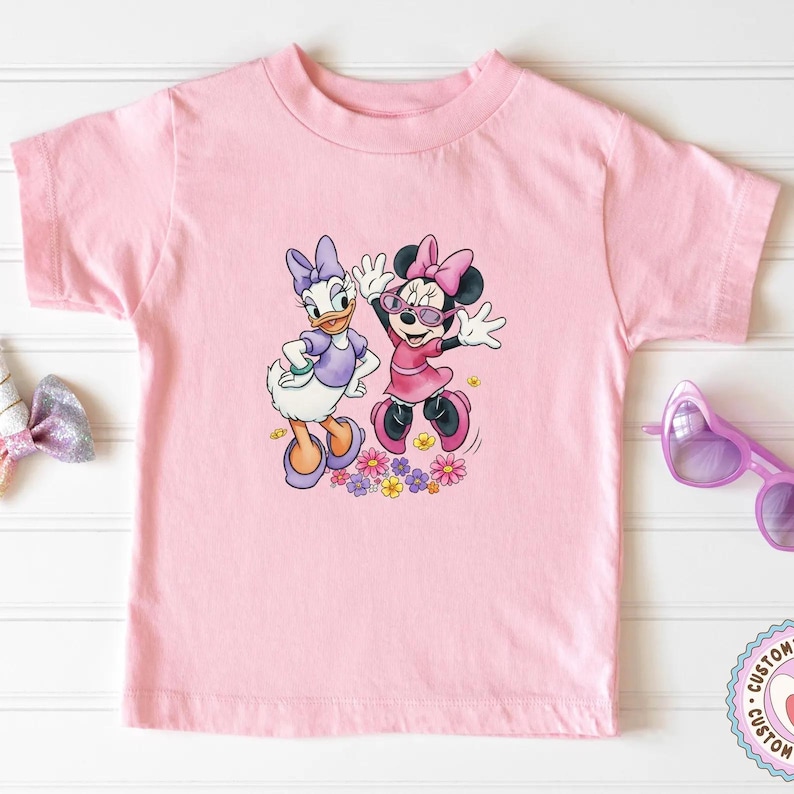 May include: A light pink t-shirt featuring a colorful graphic of Daisy Duck and Minnie Mouse. Daisy Duck is wearing a purple dress and Minnie Mouse is wearing a pink dress and sunglasses. The graphic includes colorful flowers.