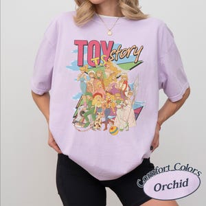 May include: A light purple short-sleeve t-shirt featuring a graphic print of various animated female characters. The text "TOY story" is printed in large letters above the characters. The design includes a colorful ball and a camera illustration.