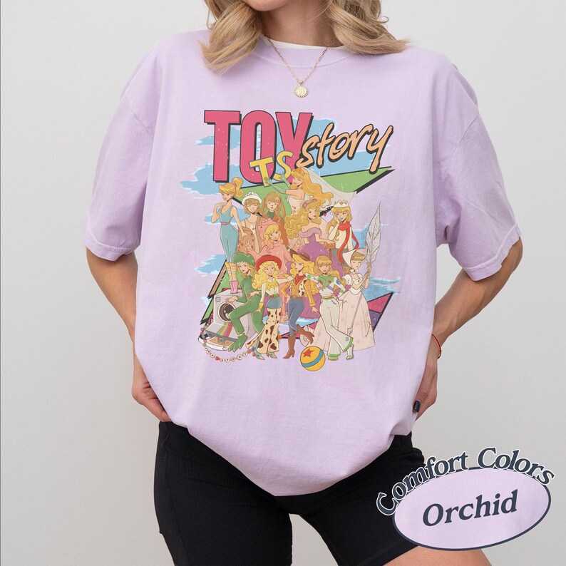 May include: A light purple short-sleeve t-shirt featuring a graphic print of various animated female characters. The text "TOY story" is printed in large letters above the characters. The design includes a colorful ball and a camera illustration.