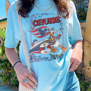 Może przedstawiać: Jasnoniebieska koszulka z grafiką Kaczora Donalda na statku wycieczkowym. Projekt zawiera napis "Let's Cruise" i "Disney Treasure", a także uszy Myszki Miki i koło ratunkowe.