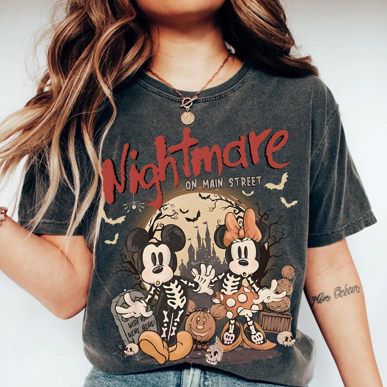 May include: Dark gray t-shirt with the words "Nightmare on Main Street" in red. The shirt features a Halloween-themed graphic of Mickey and Minnie Mouse as skeletons in front of a full moon, pumpkins, and bats.