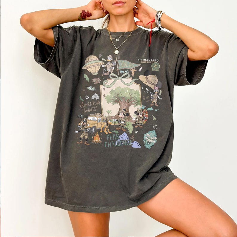 May include: Dark gray t-shirt featuring a colorful graphic with the text "Adventure Awaits!" and "Peak Challenge." The design includes cartoon characters, a hot air balloon, a jeep, and a tree. The shirt has short sleeves.