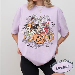 May include: A light purple short-sleeve t-shirt featuring a graphic of Disney characters in Halloween costumes, including Mickey Mouse in a pumpkin, Minnie Mouse, Goofy, and Donald Duck. The text includes names like Mickey Mouse, Minnie Mouse, and Goofy.