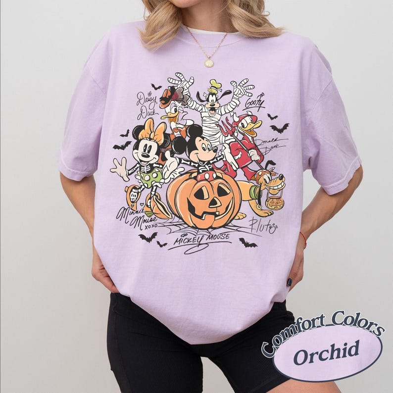 May include: A light purple short-sleeve t-shirt featuring a graphic of Disney characters in Halloween costumes, including Mickey Mouse in a pumpkin, Minnie Mouse, Goofy, and Donald Duck. The text includes names like Mickey Mouse, Minnie Mouse, and Goofy.