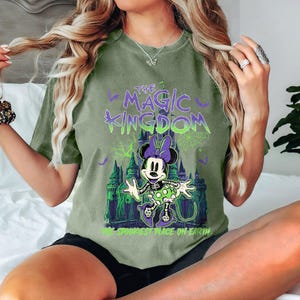 May include: Olive green t-shirt featuring a graphic of Minnie Mouse in a Halloween theme, with a skeleton design and a castle backdrop. The text reads "The Magic Kingdom" and "The Spookiest Place on Earth" in purple and green.