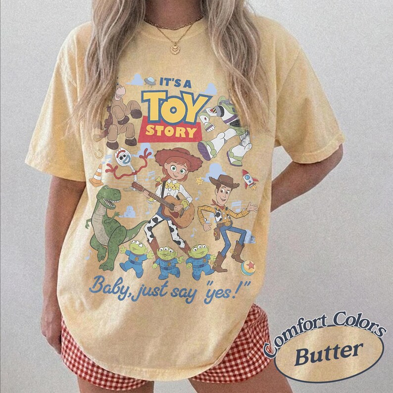 May include: A butter yellow short-sleeve t-shirt featuring a graphic print of Toy Story characters including Woody, Jessie, Rex, and the aliens. The text reads IT'S A TOY STORY and Baby, just say yes! The shirt has a crew neckline and a relaxed fit.