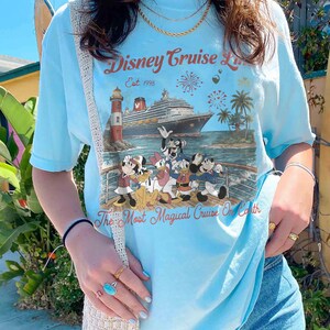 May include: Pale blue t-shirt with a Disney Cruise Line graphic. The design includes a cruise ship, lighthouse, palm trees, and Disney characters. Text reads "Disney Cruise Line Est. 1998" and "The Most Magical Cruise On Earth".