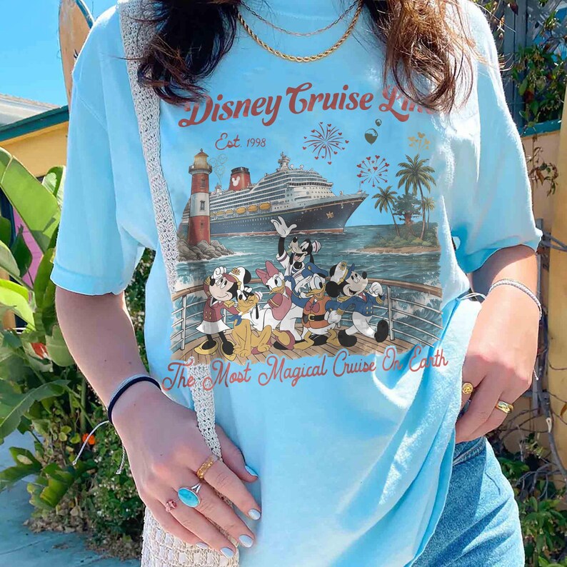 May include: Pale blue t-shirt with a Disney Cruise Line graphic. The design includes a cruise ship, lighthouse, palm trees, and Disney characters. Text reads "Disney Cruise Line Est. 1998" and "The Most Magical Cruise On Earth".