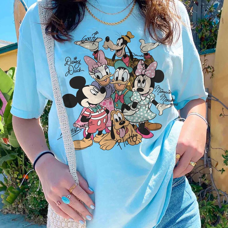 May include: Light blue t-shirt featuring a graphic of Disney characters including Mickey Mouse, Minnie Mouse, Donald Duck, Daisy Duck, Goofy, and Pluto. The shirt has the text "Mickey Mouse" and "Minnie Mouse" on it.