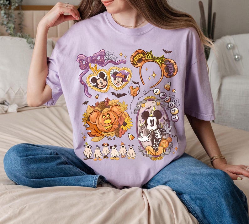 May include: A light purple t-shirt featuring a Halloween-themed graphic. The design includes Mickey and Minnie Mouse, a pumpkin, ghosts, and the text "Spooky". The shirt has a relaxed fit and short sleeves.