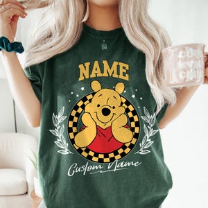 K&ouml;nnte beinhalten: Dunkelgr&uuml;nes T-Shirt mit einem Cartoon-B&auml;ren-Design. Das Wort "NAME" steht &uuml;ber dem B&auml;ren und "Custom Name" darunter. Der B&auml;r befindet sich in einem schwarz-gelb karierten Kreis mit wei&szlig;en Sternen und Bl&auml;ttern. Der B&auml;r tr&auml;gt ein rotes Hemd.