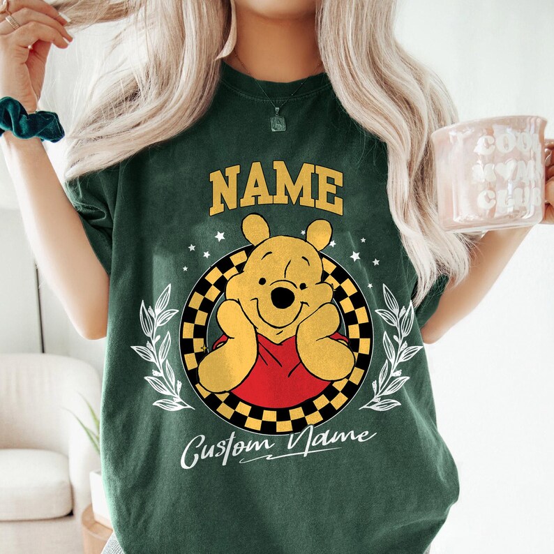 K&ouml;nnte beinhalten: Dunkelgr&uuml;nes T-Shirt mit einem Cartoon-B&auml;ren-Design. Das Wort "NAME" steht &uuml;ber dem B&auml;ren und "Custom Name" darunter. Der B&auml;r befindet sich in einem schwarz-gelb karierten Kreis mit wei&szlig;en Sternen und Bl&auml;ttern. Der B&auml;r tr&auml;gt ein rotes Hemd.