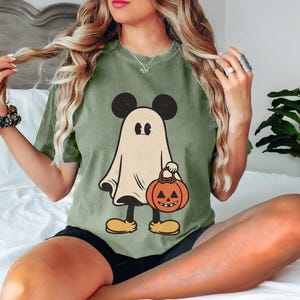 May include: Green t-shirt with a cartoon ghost wearing a Mickey Mouse hat and holding a jack-o'-lantern. The ghost is white with black eyes and a black mouth.