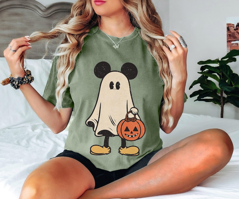 May include: Green t-shirt with a cartoon ghost wearing a Mickey Mouse hat and holding a jack-o'-lantern. The ghost is white with black eyes and a black mouth.