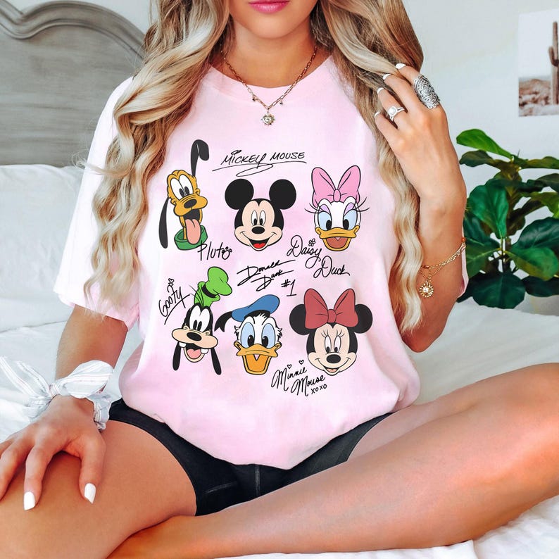 May include: Pink t-shirt with a graphic print of Mickey Mouse, Minnie Mouse, Donald Duck, Daisy Duck, Goofy, and Pluto. The shirt has the text "Mickey Mouse" and "Minnie Mouse xoxo" printed on it.