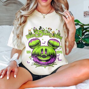 May include: Cream-colored t-shirt featuring a Halloween-themed graphic. The design includes a green and purple Mickey Mouse pumpkin, the text "Walt Disney World" and "Est. 1971". Small ghosts and bats are also included in the design.
