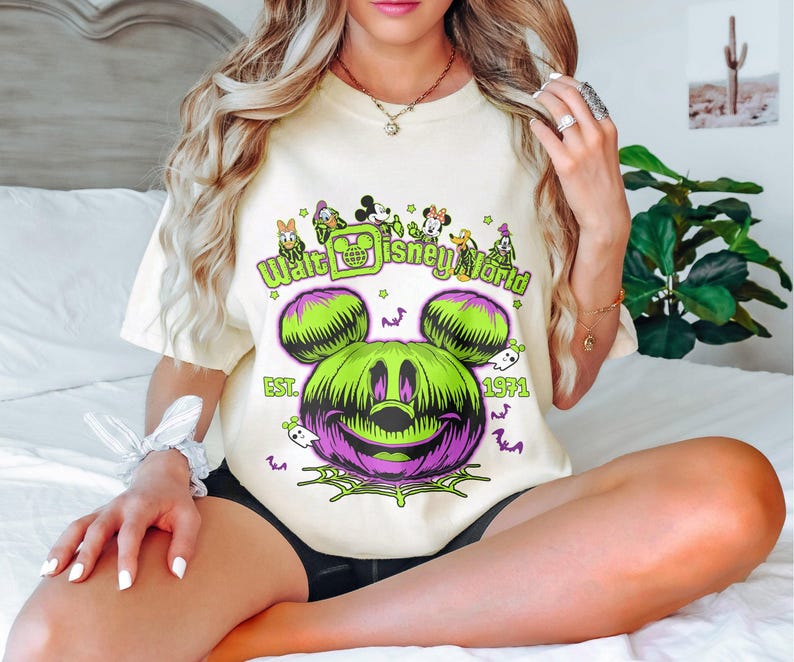 May include: Cream-colored t-shirt featuring a Halloween-themed graphic. The design includes a green and purple Mickey Mouse pumpkin, the text "Walt Disney World" and "Est. 1971". Small ghosts and bats are also included in the design.