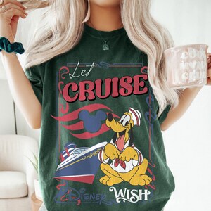 Może przedstawiać: Ciemnozielona koszulka z grafiką z napisem "Let Cruise" w kolorze czerwonym i białym, postacią z kresk&oacute;wki Pluto i statkiem wycieczkowym. Koszulka zawiera r&oacute;wnież napis "Disney Wish".