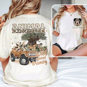 May include: Cream-colored t-shirt featuring a graphic of Mickey Mouse and friends in a brown jeep with the text "Animal Kingdom" and "Wild about Disney". The shirt also has a small Mickey Mouse graphic on the front.