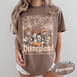 May include: Espresso-colored t-shirt featuring a Halloween-themed Disneyland graphic. The design includes cartoon characters in skeleton costumes, a haunted castle, and the text "Disneyland Est. 1955". The shirt is from Comfort Colors.