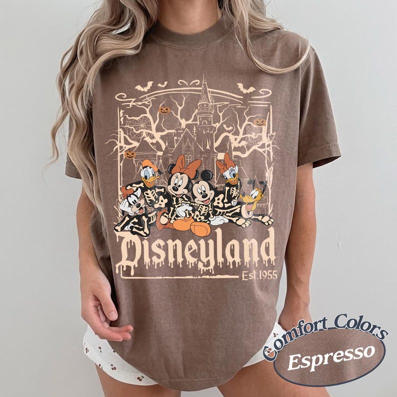 May include: Espresso-colored t-shirt featuring a Halloween-themed Disneyland graphic. The design includes cartoon characters in skeleton costumes, a haunted castle, and the text "Disneyland Est. 1955". The shirt is from Comfort Colors.