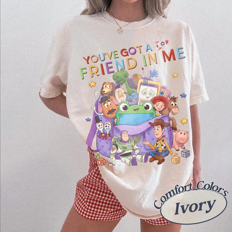 May include: An ivory t-shirt featuring the text "YOU'VE GOT A FRIEND IN ME" above a graphic of various animated characters, including a cowboy, a space ranger, a dinosaur, a potato figure, and a green frog, all emerging from a purple backpack.
