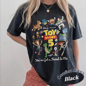 May include: A black acid-wash t-shirt featuring a colorful graphic of Disney Pixar Toy Story characters including Woody, Buzz Lightyear, and Rex. The design includes the text Toy Story 5 and You're Got a Friend In Me, with a blank space for a name.