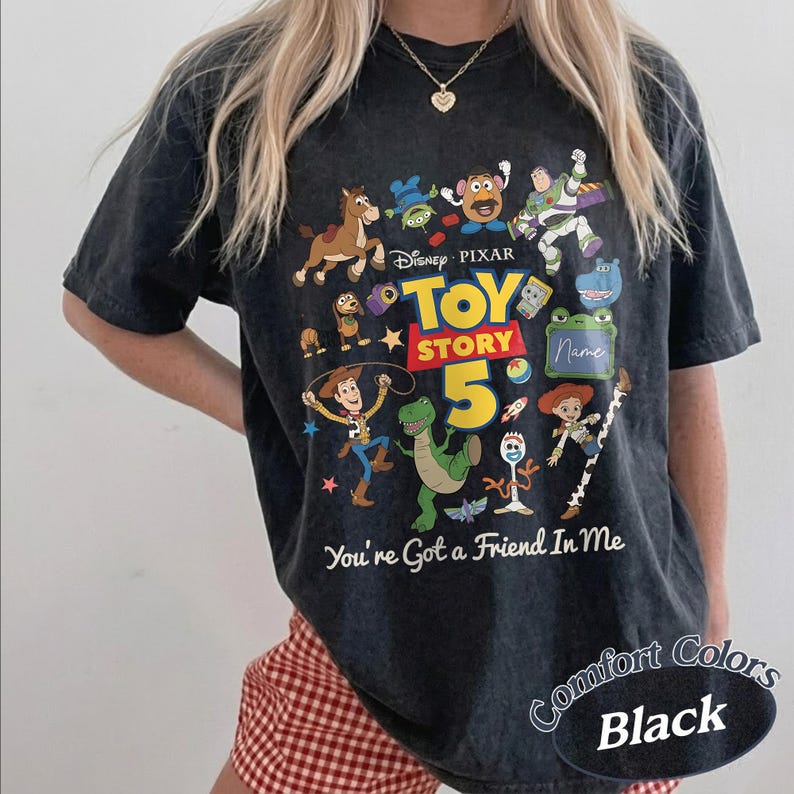 May include: A black acid-wash t-shirt featuring a colorful graphic of Disney Pixar Toy Story characters including Woody, Buzz Lightyear, and Rex. The design includes the text Toy Story 5 and You're Got a Friend In Me, with a blank space for a name.