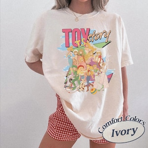 May include: An ivory short-sleeve t-shirt featuring a graphic print with the text "TOY story" in pink and blue. The design displays a group of animated characters in various outfits, including a cowboy hat and boots, set against a geometric background.
