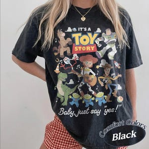 May include: A black short-sleeve t-shirt featuring a graphic print of Toy Story characters including Woody, Buzz Lightyear, Jessie, Bullseye, Rex, Forky, and the aliens. The text reads IT'S A TOY STORY and Baby, just say yes! in white and yellow.