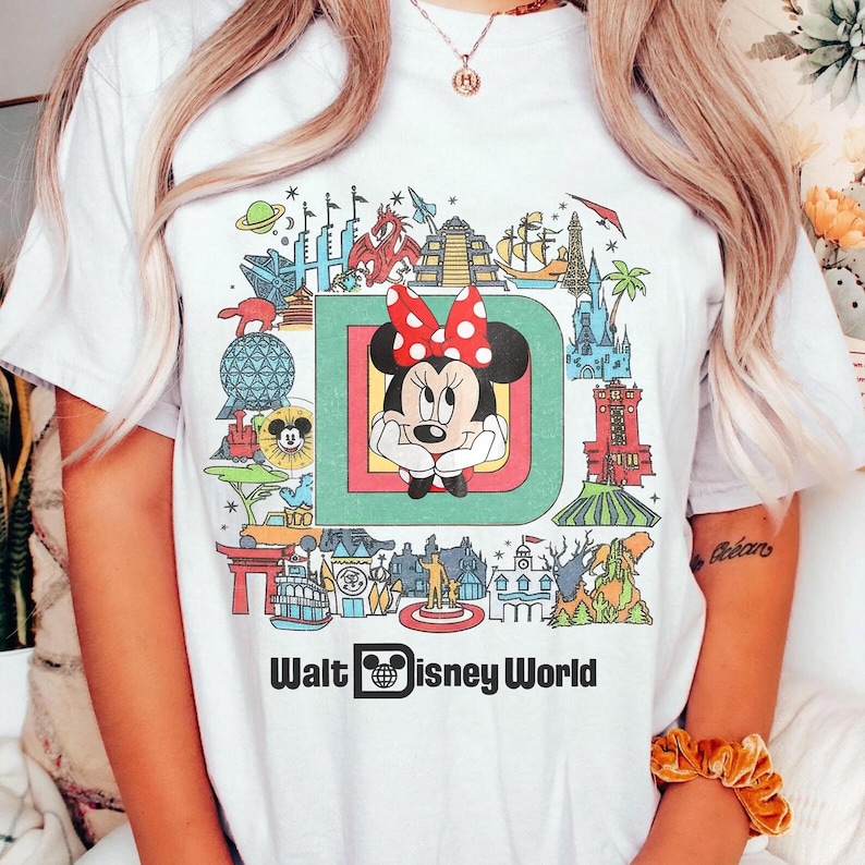 Puede incluir: Camiseta blanca con un gr&aacute;fico colorido de Minnie Mouse en un marco verde y rojo, rodeada de iconos de Disney World. Las palabras "Walt Disney World" est&aacute;n impresas debajo.
