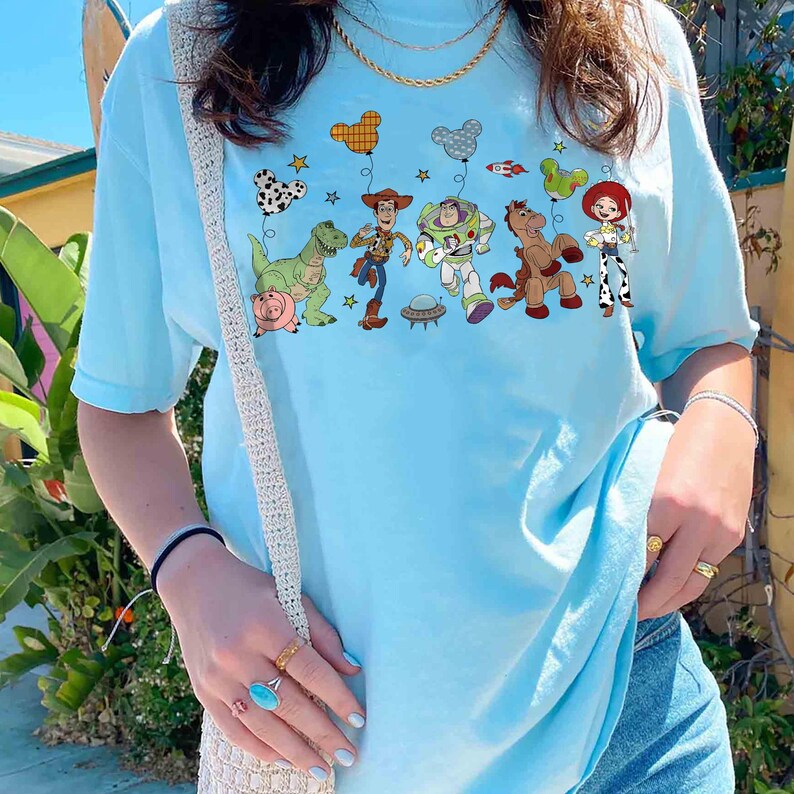 Puede incluir: Camiseta azul claro con un gr&aacute;fico colorido de personajes animados, incluyendo un dinosaurio y un vaquero. El dise&ntilde;o incluye globos y estrellas. La camiseta est&aacute; hecha de un material suave.
