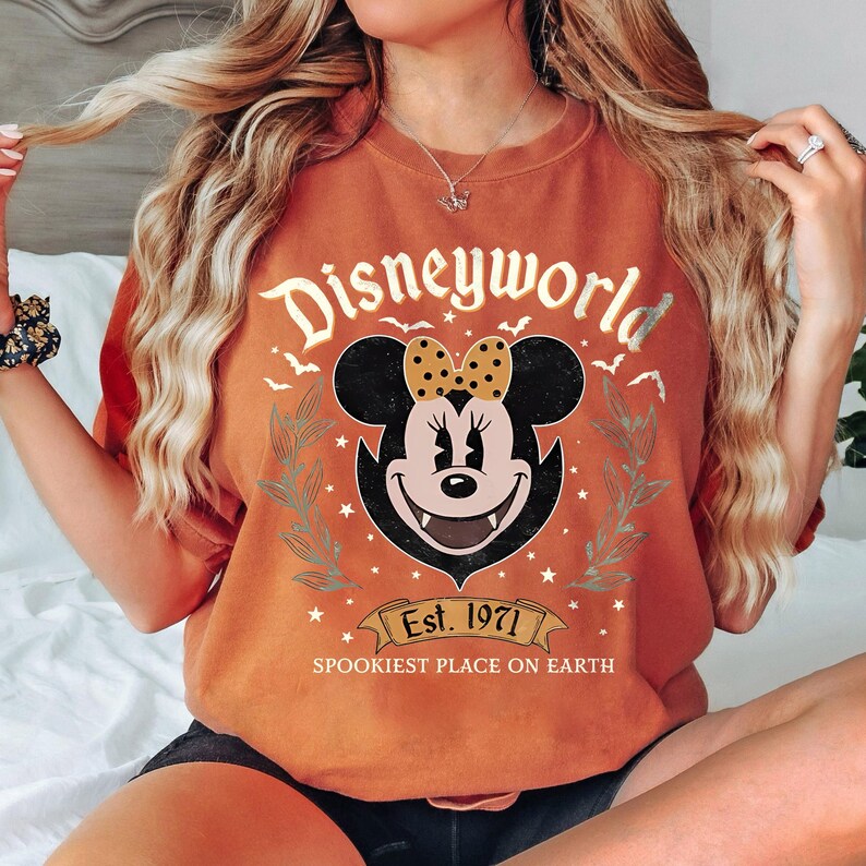 May include: Orange t-shirt with a graphic of Minnie Mouse as a vampire, with the text "Disneyworld" above her head. The shirt also features the text "Est. 1971" and "Spookiest Place on Earth" on a banner below Minnie.