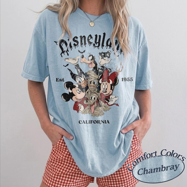 May include: A light blue short-sleeve t-shirt featuring a graphic print of Disney characters dressed in Halloween costumes. The text reads Disneyland, Est 1955, and California. The design includes Mickey, Minnie, Goofy, Donald, and Pluto.