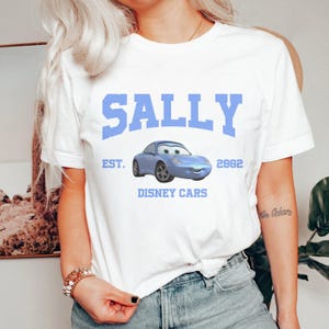Puede incluir: Camiseta blanca con un gr&aacute;fico azul de Sally de Cars de Disney. El gr&aacute;fico incluye el texto "Sally", "Est. 2002", y "Disney Cars".