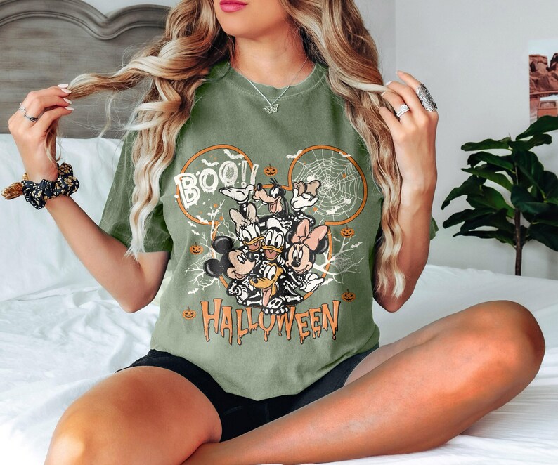 K&ouml;nnte beinhalten: Olivgr&uuml;nes T-Shirt mit einem Halloween-Motiv. Das Design zeigt Micky Maus, Donald Duck, Goofy und Minnie Maus, mit den Worten "BOO!!" und "HALLOWEEN" in Orange. Die Grafik zeigt auch ein Spinnennetz und K&uuml;rbisse.