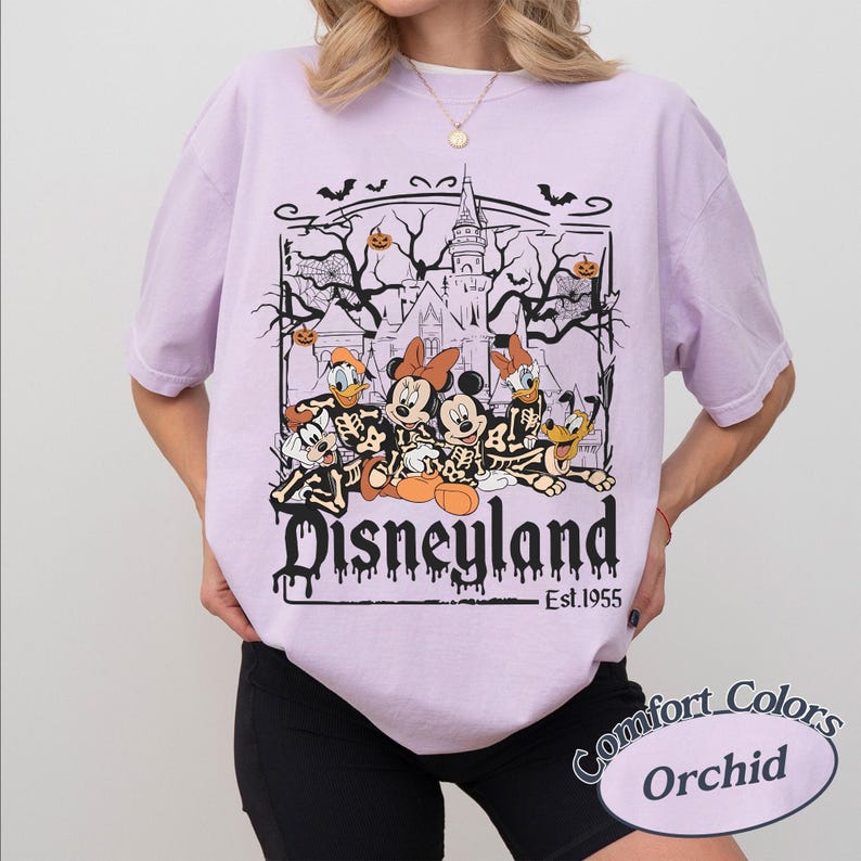 May include: Orchid-colored t-shirt featuring a Halloween-themed Disneyland graphic. The design includes cartoon characters in skeleton costumes, a haunted castle, bats, and the text "Disneyland Est. 1955". The shirt is a Comfort Colors brand.
