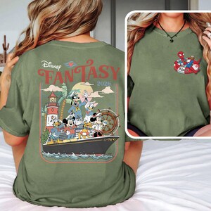 May include: An olive green t-shirt with a Disney Fantasy 2026 design on the back. The graphic shows Mickey Mouse, Minnie Mouse, Donald Duck, Goofy, and other characters on a cruise ship. A smaller graphic of Spider-Man and Santa Claus is on the front.